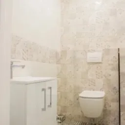 Nowoczesna, jasna łazienka z wiszącą toaletą, białą szafką pod umywalką z chromowanymi uchwytami i wzorzystymi płytkami podłogowymi oraz ściennymi w odcieniach szarości i beżu.