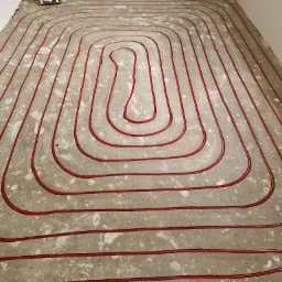 Czerwone rurki ogrzewania podłogowego ułożone spiralnie na szarym, surowym betonie posadzki w trakcie instalacji.