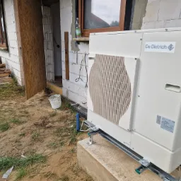 Zewnętrzna jednostka pompy ciepła marki De Dietrich, zamontowana na betonowym fundamencie przy budynku w trakcie budowy, widoczna butelka z wodą na parapecie okna.