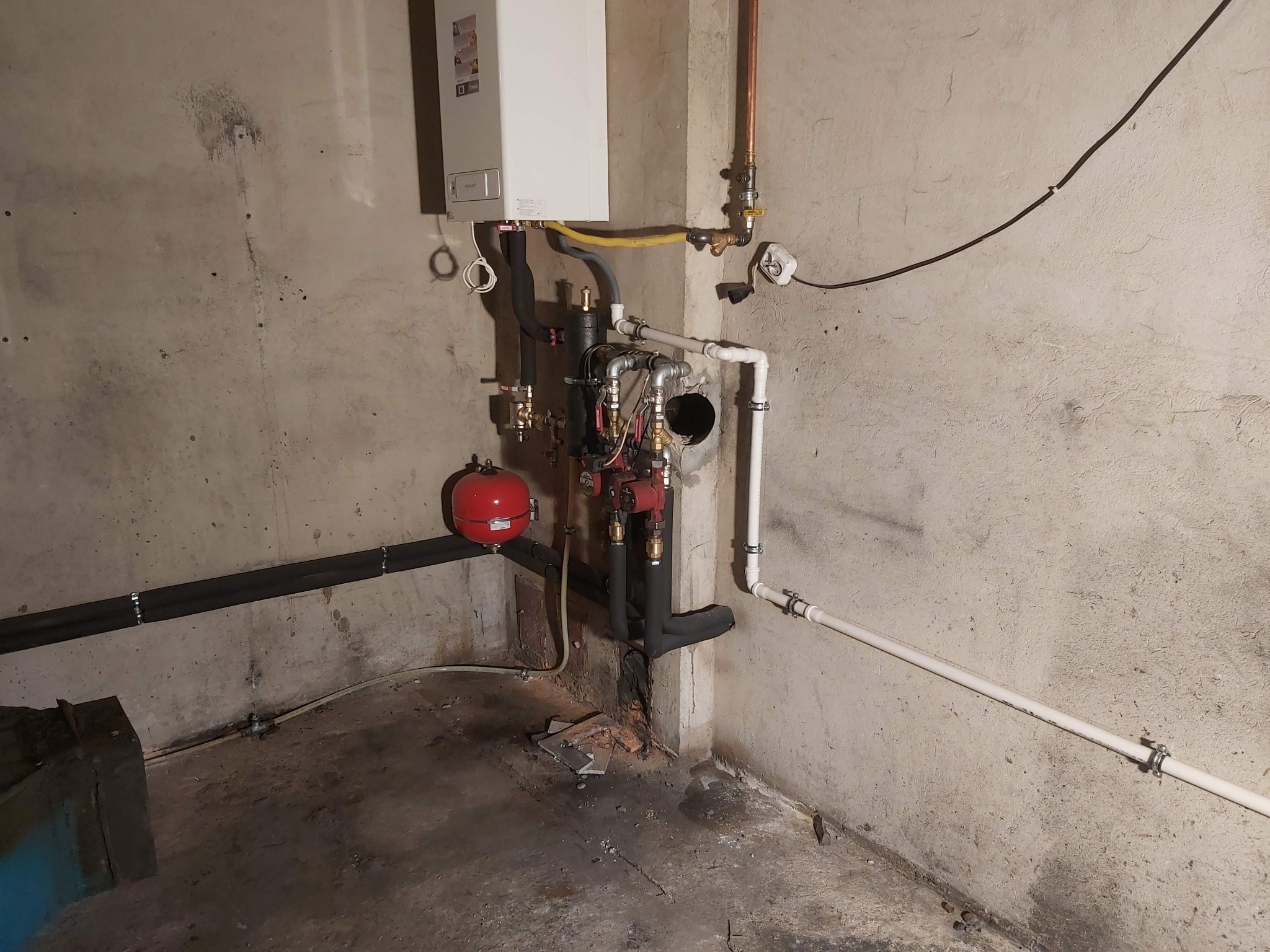 Kocioł gazowy wiszący na szarej, surowej ścianie piwnicy, z widocznymi rurami miedzianymi i plastikowymi, zbiornikiem wyrównawczym i pompą obiegową. Na podłodze widoczne resztki gruzu i pyłu.