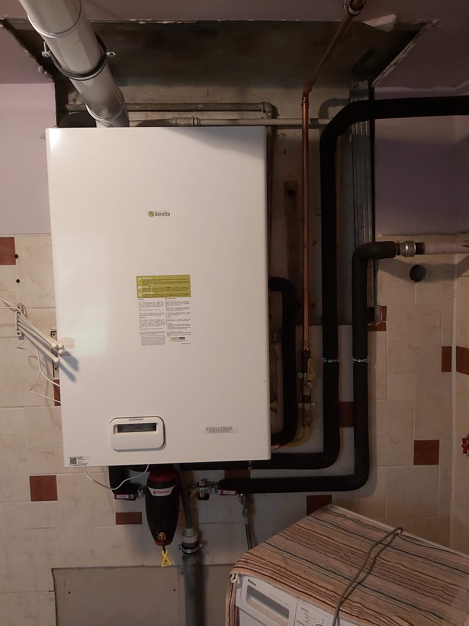 Biały wiszący piec gazowy Beretta z podłączonymi rurami miedzianymi i czarnymi izolowanymi. Filtr magnetyczny Flamco pod kotłem. Widoczna pralka Amica z narzutą w tle.