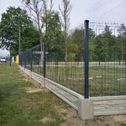 Nowoczesne ogrodzenie z paneli z podmurówką imitującą kamień, widok z boku na tle zieleni i drzew