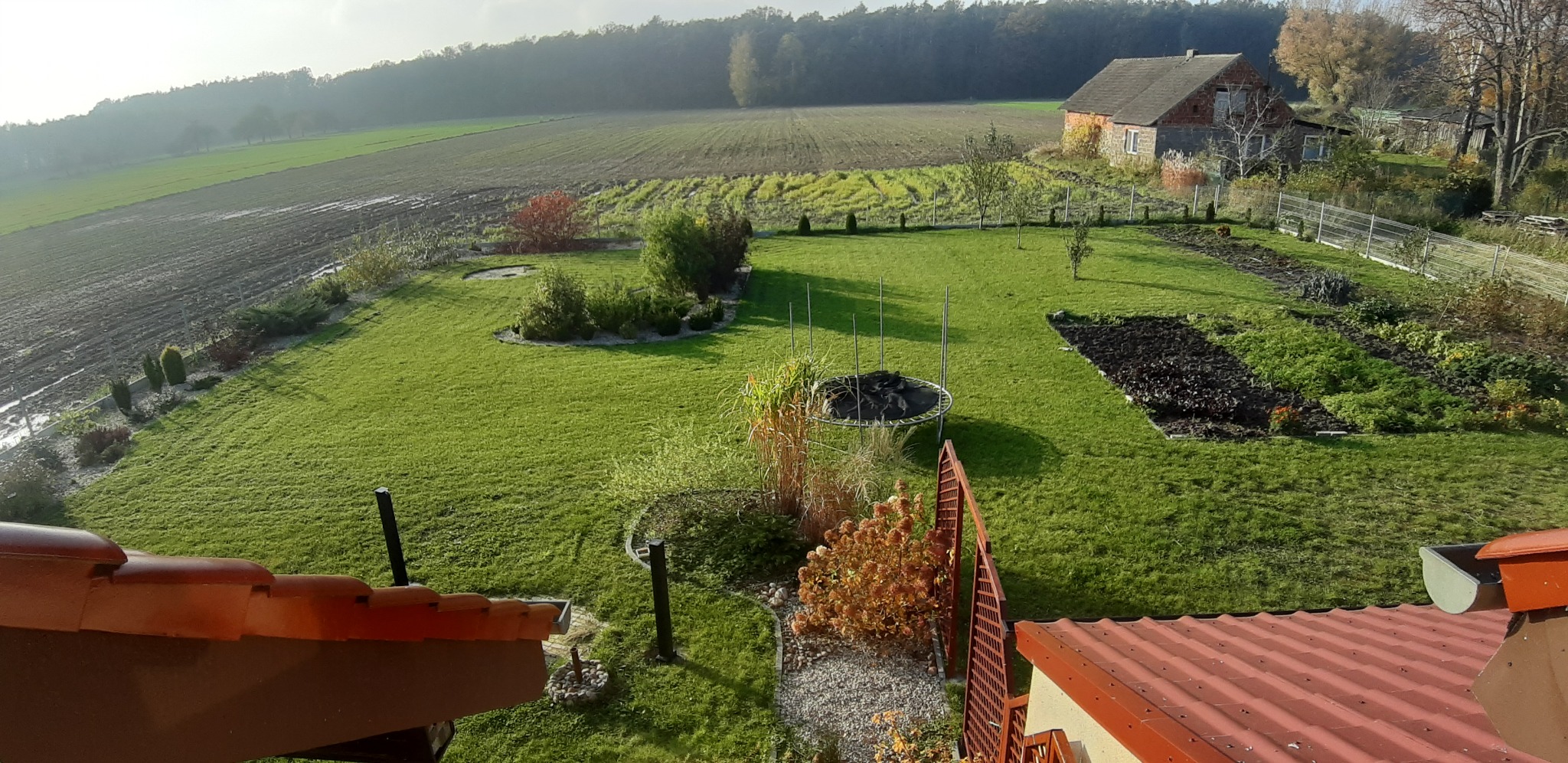Widok z góry na duży, zadbany ogród z trawnikiem, rabatami kwiatowymi, trampoliną i domem w tle, w słoneczny dzień.