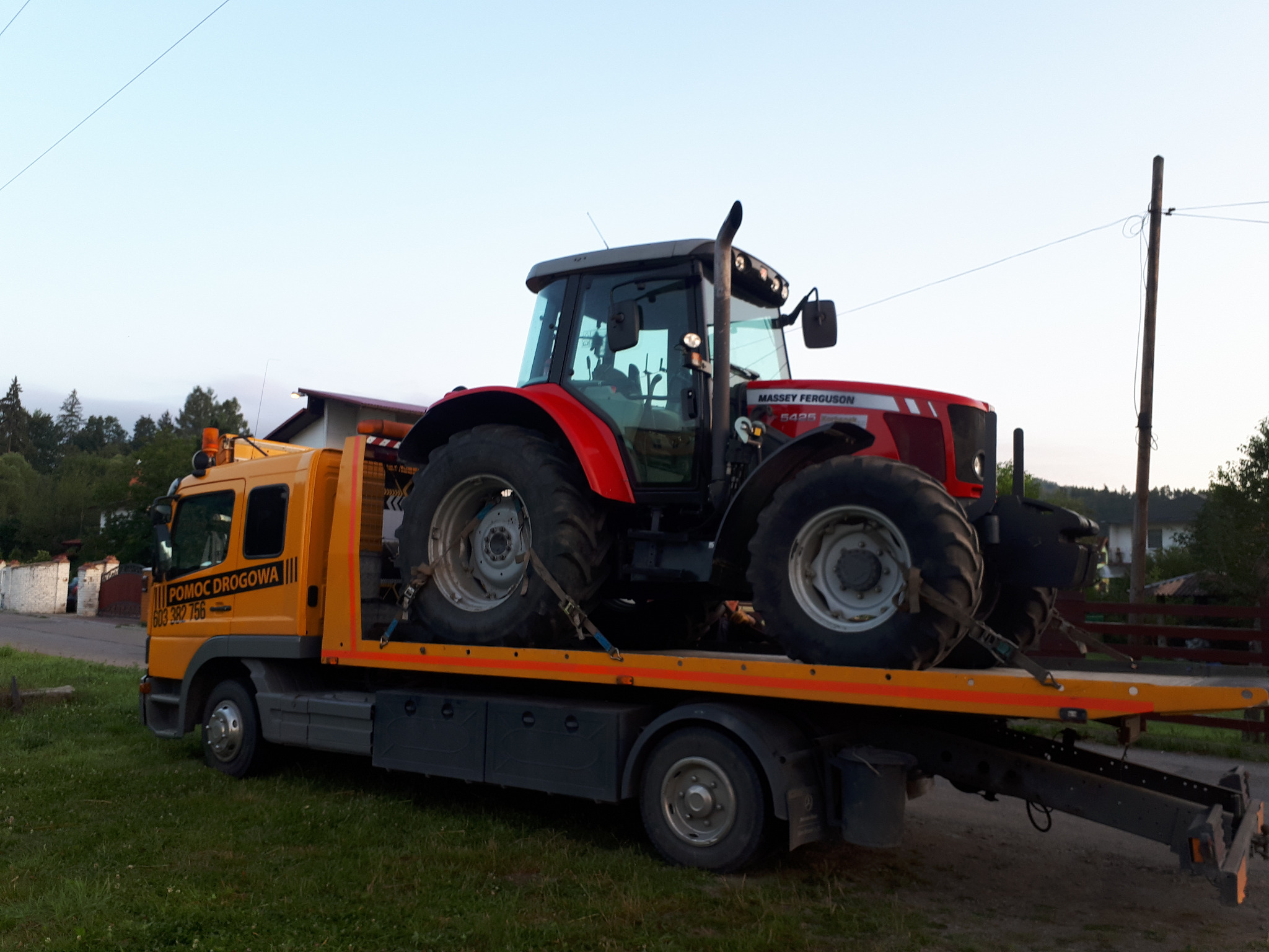 Żółta laweta 'Pomoc Drogowa' przewozi czerwony traktor Massey Ferguson 5425, zabezpieczony pasami transportowymi.