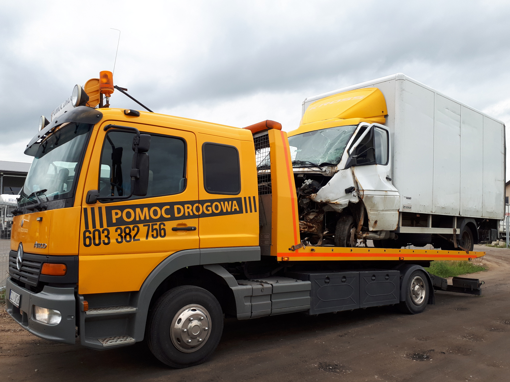 Żółty samochód pomocy drogowej Mercedes-Benz z uszkodzonym białym furgonem na platformie, widoczne numery telefonu alarmowego i zniszczony przód furgonu