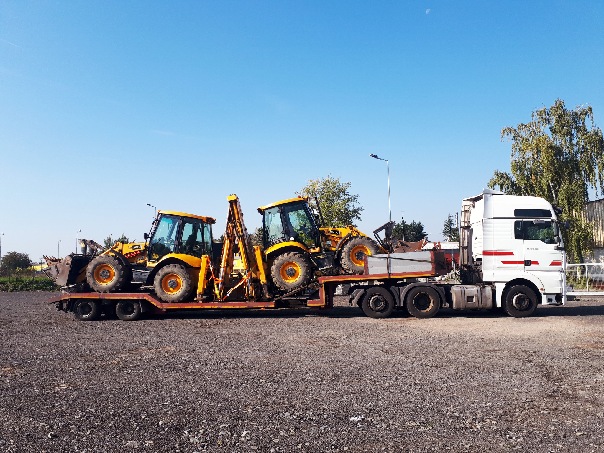 Ciężarówka z naczepą przewozi dwie żółte koparko-ładowarki JCB 3CX na tle błękitnego nieba z widocznym księżycem.