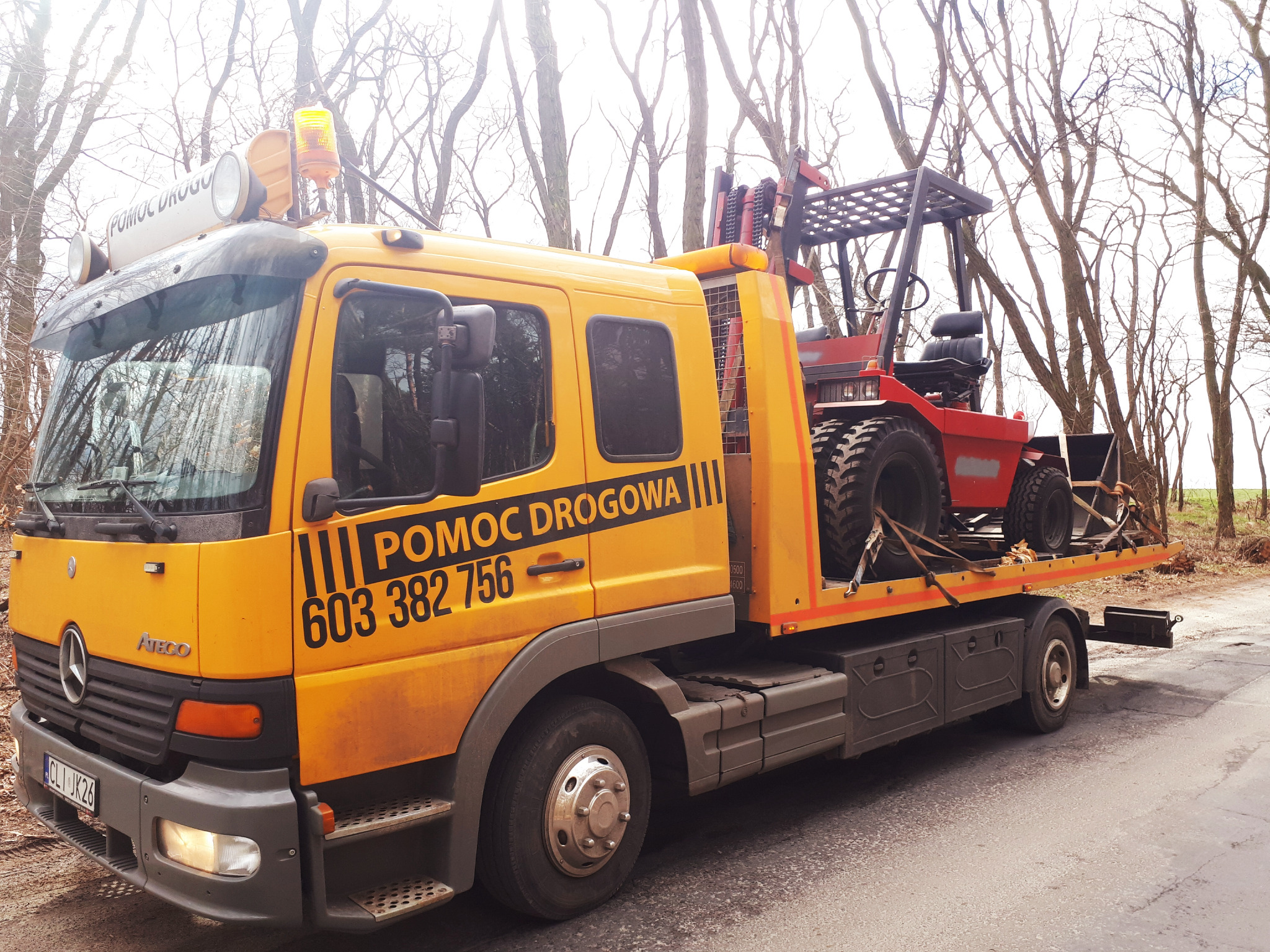 Żółta laweta Mercedes-Benz Atego z napisem 'Pomoc Drogowa' przewożąca czerwony wózek widłowy na platformie załadunkowej, na tle drzew bez liści.