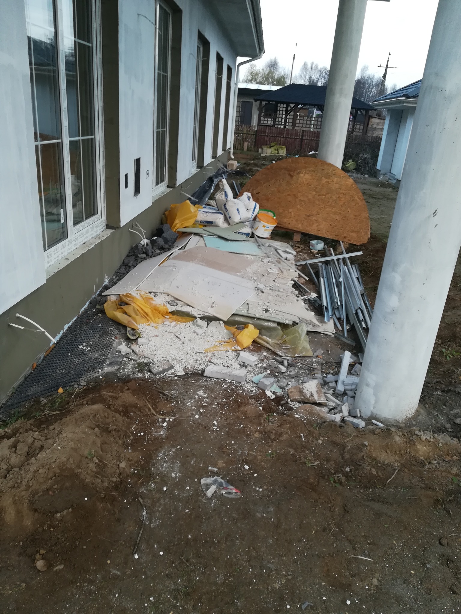 Teren wokół domu w trakcie prac budowlanych: widoczne fundamenty, okna, materiały budowlane, ziemia i gruz.