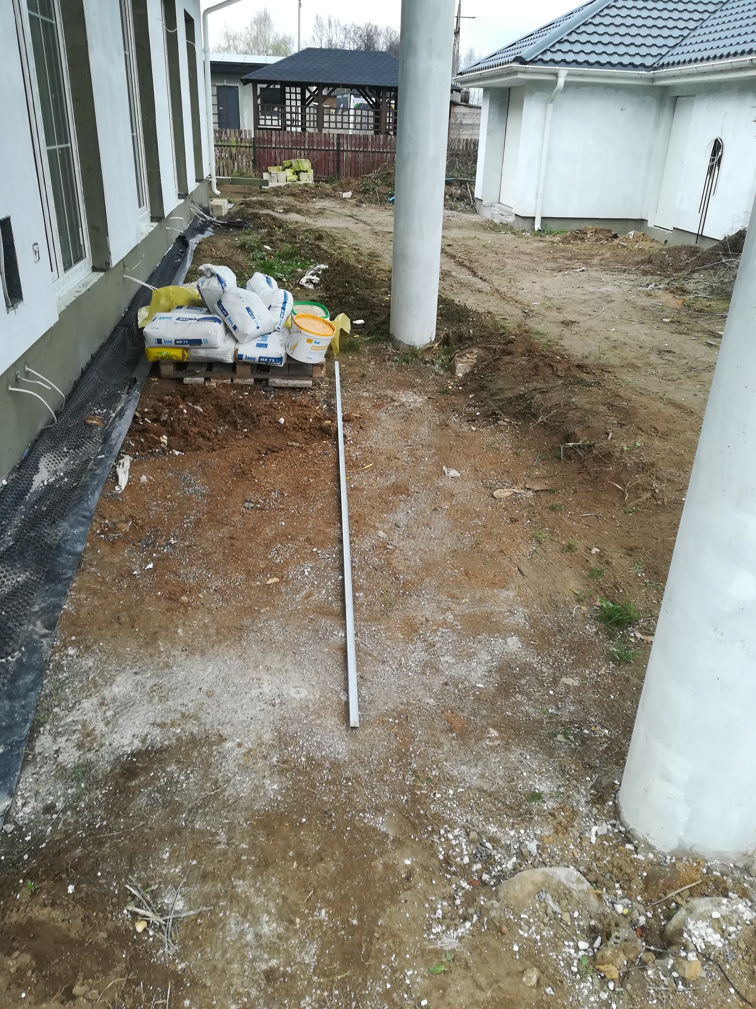 Przygotowanie terenu pod przyszły ogród: worki z zaprawą, wiaderka i rura leżą na ziemi obok budynku z oknami i kolumnami.