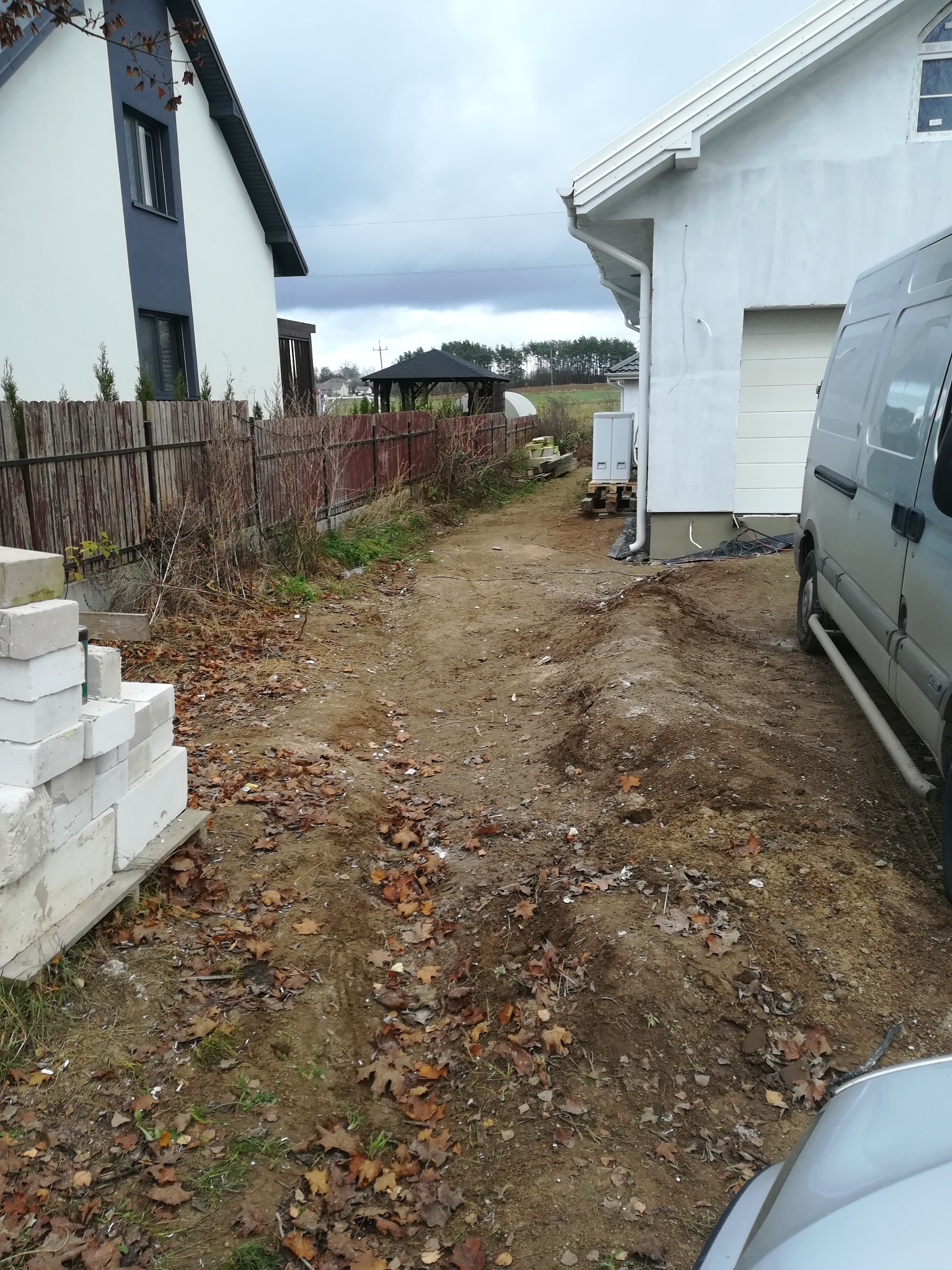 Przygotowanie terenu pod ogród w Łomży: wyrównana ziemia, sterta białych bloczków na palecie, częściowo widoczny bus i dwa budynki w tle, jesienne liście na ziemi.