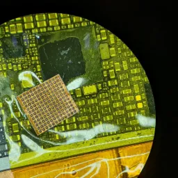 Zbliżenie pod mikroskopem na płytę główną urządzenia elektronicznego z widocznym układem BGA (Ball Grid Array) i śladami topnika lutowniczego.