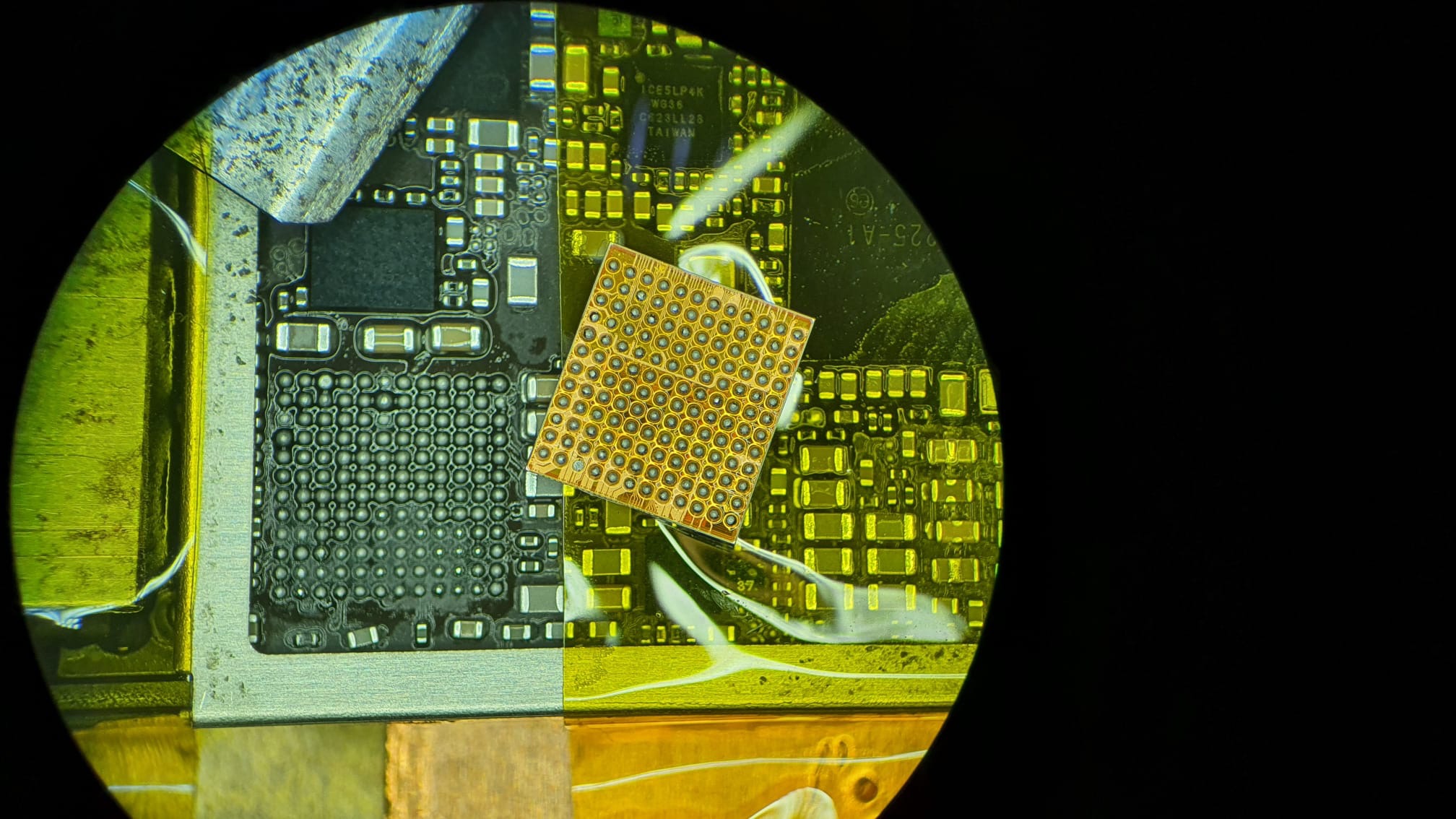 Zbliżenie na płytę główną telefonu pod mikroskopem, widoczne układy scalone, elementy elektroniczne i złote punkty lutownicze.