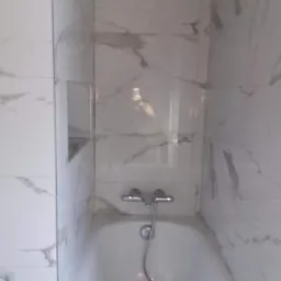 Wykończona łazienka z wanną i ścianami wyłożonymi białymi płytkami imitującymi marmur, widoczna wnęka w ścianie.