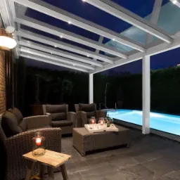 Metalowa pergola z oświetleniem LED nad tarasem z meblami ogrodowymi i basenem, widok nocą.
