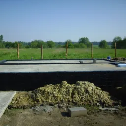 Betonowa płyta fundamentowa z izolacją przeciwwilgociową, widok na teren budowy w słoneczny dzień, w tle zielone pola i drzewa.