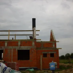 Surowy stan domu z czerwonej cegły w trakcie budowy, widoczne niezabezpieczone otwory okienne i komin, na pierwszym planie materiały budowlane i betoniarka, pochmurne niebo w tle.