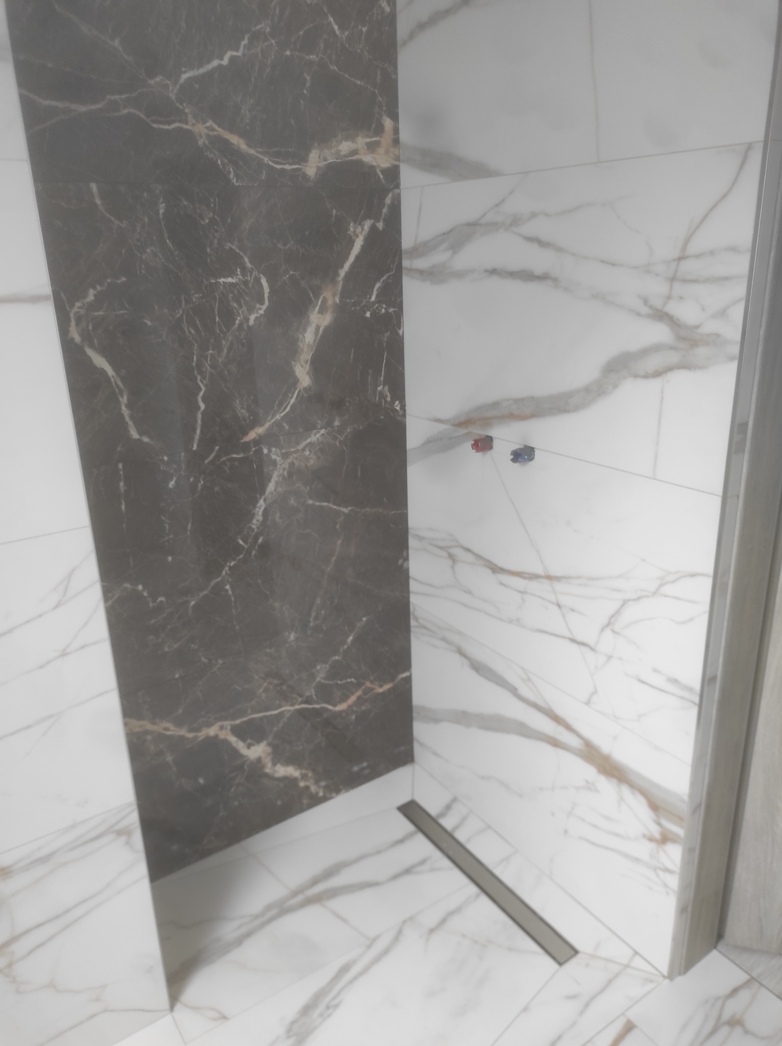 Wykończona ściana i podłoga w łazience z białych i grafitowych płytek imitujących marmur, widoczny odpływ liniowy i fragment kabiny prysznicowej.