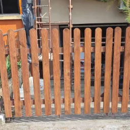 Świeżo pomalowane drewniane ogrodzenie z zaokrąglonymi sztachetami, widoczne ślady malowania na betonowej podstawie, drabina oparta o budynek w tle.