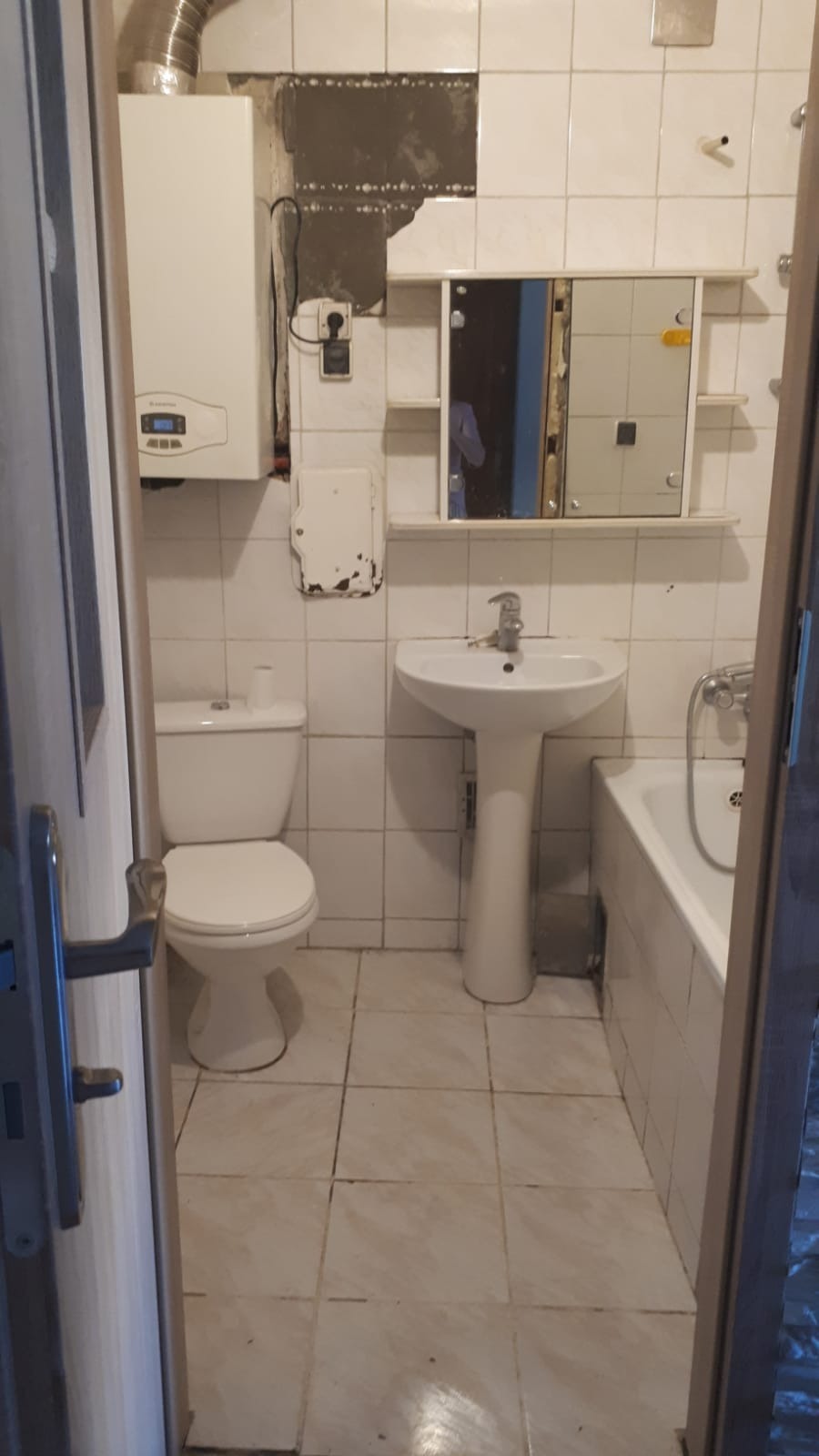 Zdjęcie łazienki w trakcie remontu: widoczne białe płytki, umywalka na postumencie, toaleta, wanna, bojler oraz fragment ściany z usuniętymi płytkami i odsłoniętym murem, sugerujące prace...