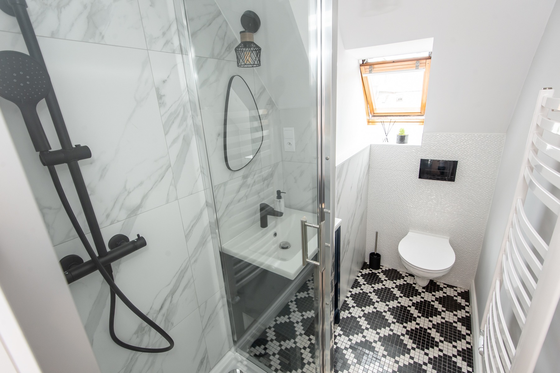 Nowoczesna łazienka z kabiną prysznicową, umywalką, toaletą i czarno-białą mozaiką podłogową, oświetlona światłem dziennym z okna dachowego.