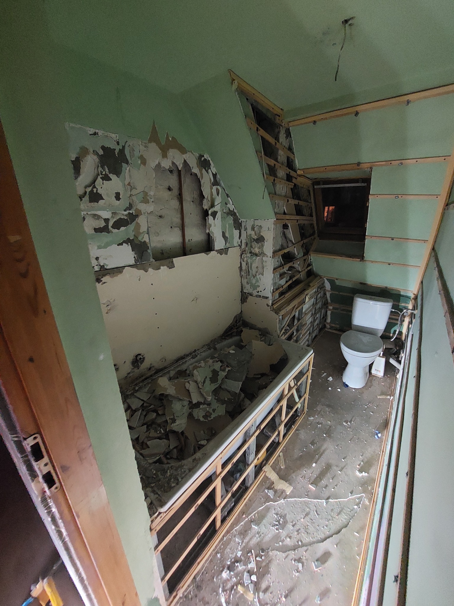 Łazienka w trakcie demontażu: odsłonięte fragmenty ścian, pozostałości płytek w wannie, widoczna konstrukcja drewniana i toaleta.