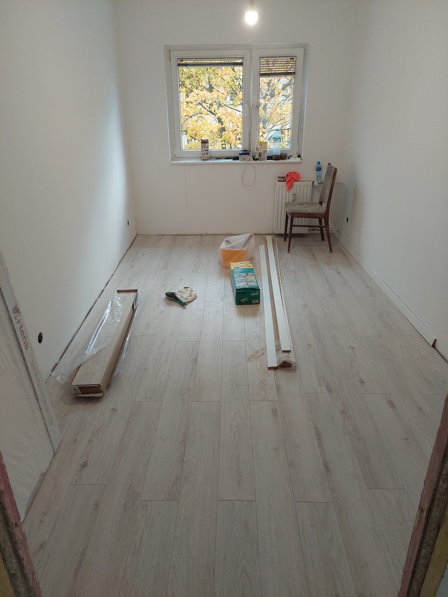Pokój z nową, jasną podłogą laminowaną, listwami przypodłogowymi i oknem z widokiem na jesienne drzewa, przygotowany do dalszych prac wykończeniowych.
