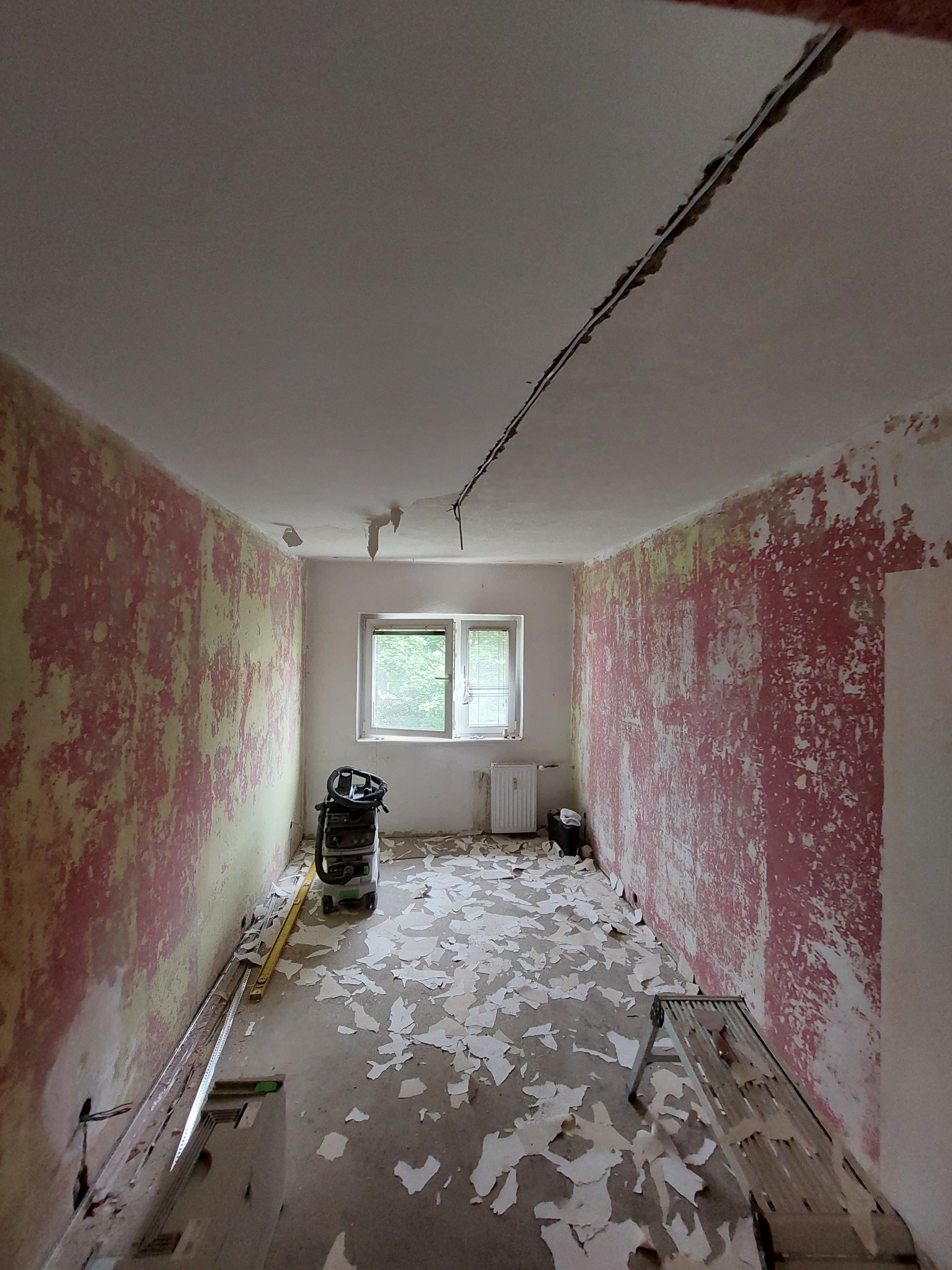 Pokój w trakcie remontu: ściany z resztkami starej farby w odcieniach różu i żółci, podłoga pokryta odpadami, widoczny odkurzacz przemysłowy i drabina, przygotowania do malowania lub szpachlowania.