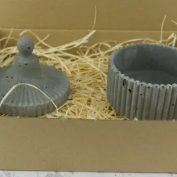 Dekoracyjny, betonowy pojemnik z pokrywką w kartonowym pudełku wypełnionym siankiem, prezentowany przez firmę z Gorzowa Wielkopolskiego.