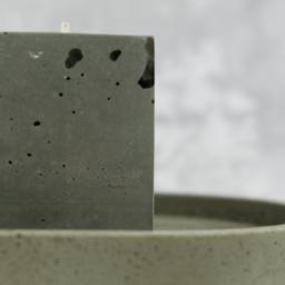 Rausch Beton - Szara, betonowa świeca w kształcie sześcianu, umieszczona na owalnej, betonowej podstawce z widocznymi porami i niedoskonałościami, na rozmytym, jasnoszarym tle.