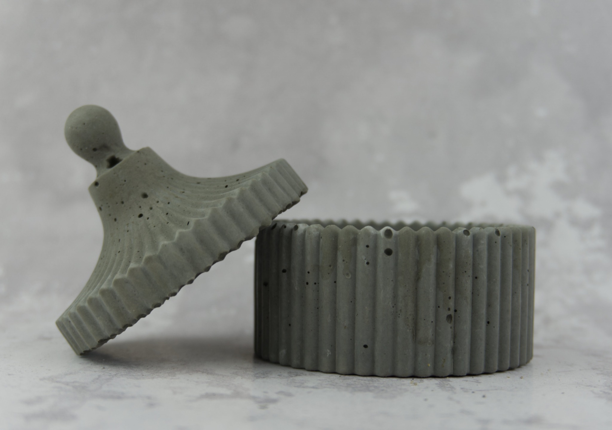 Dekoracyjny, betonowy pojemnik z pokrywką, o żłobkowanej fakturze, na tle szarego, marmurkowego blatu.