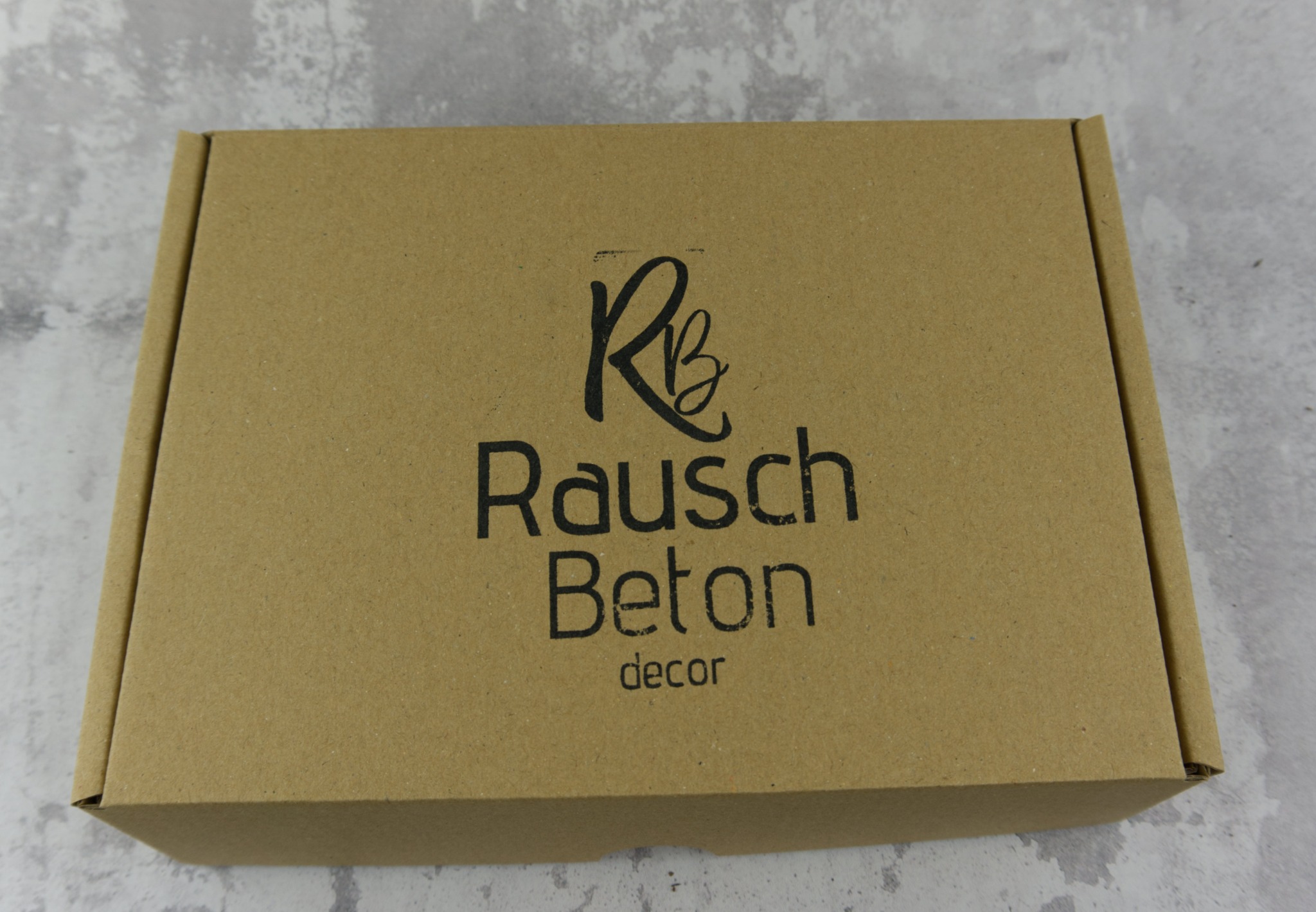 Kartonowe pudełko z czarnym logo i napisem 'Rausch Beton decor' na szarym tle.