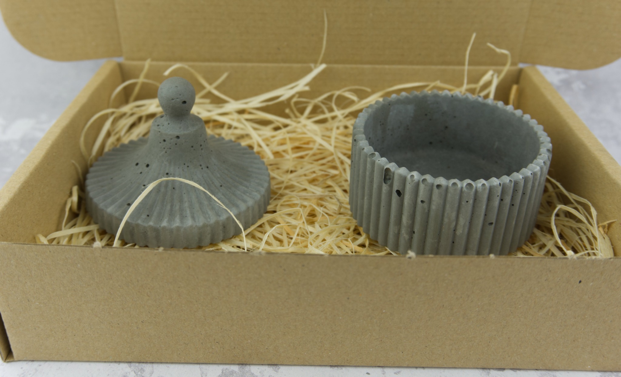 Dekoracyjny, betonowy pojemnik z pokrywką w kartonowym pudełku wypełnionym siankiem, prezentowany przez firmę z Gorzowa Wielkopolskiego.