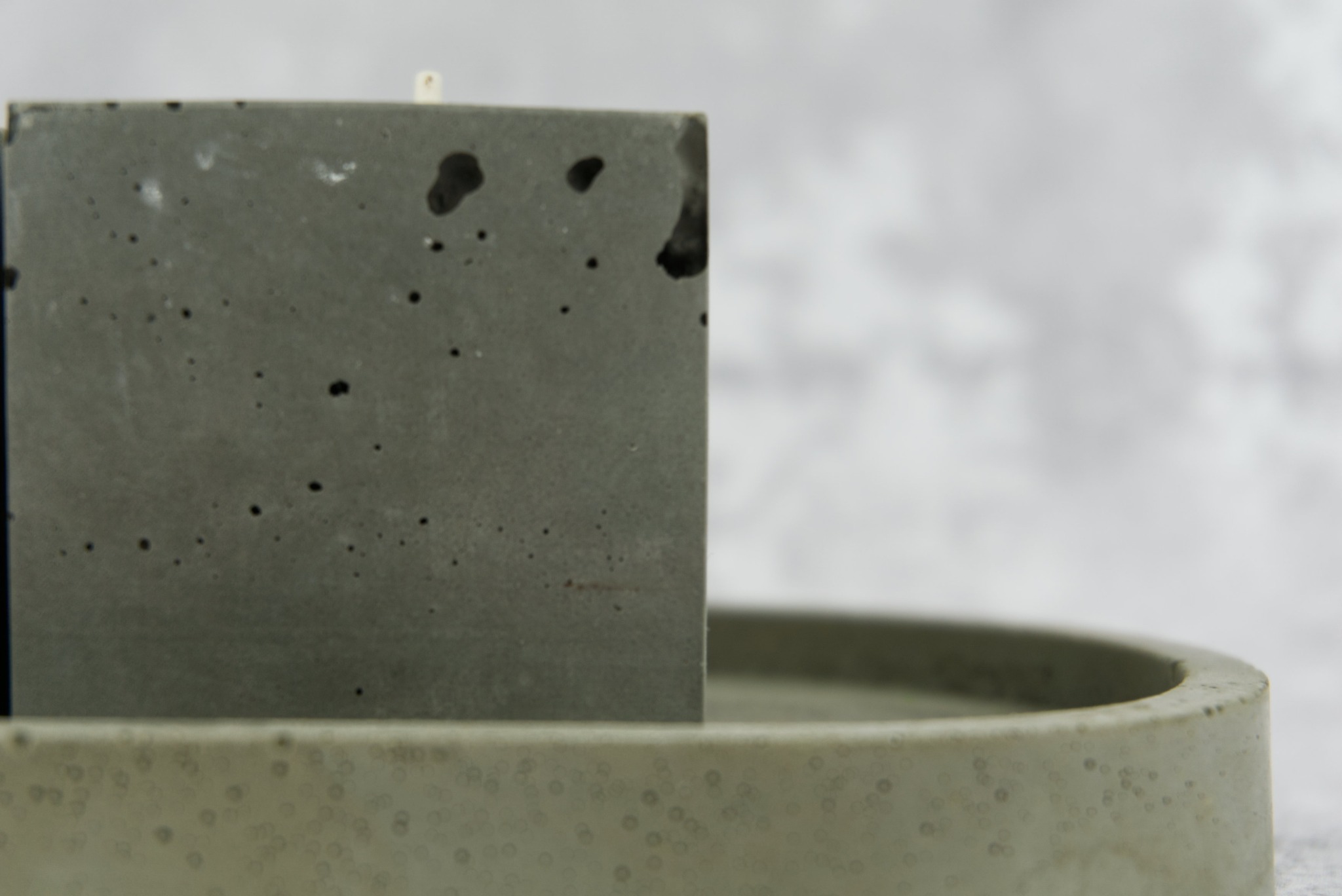 Szara, betonowa świeca w kształcie sześcianu, umieszczona na owalnej, betonowej podstawce z widocznymi porami i niedoskonałościami, na rozmytym, jasnoszarym tle.