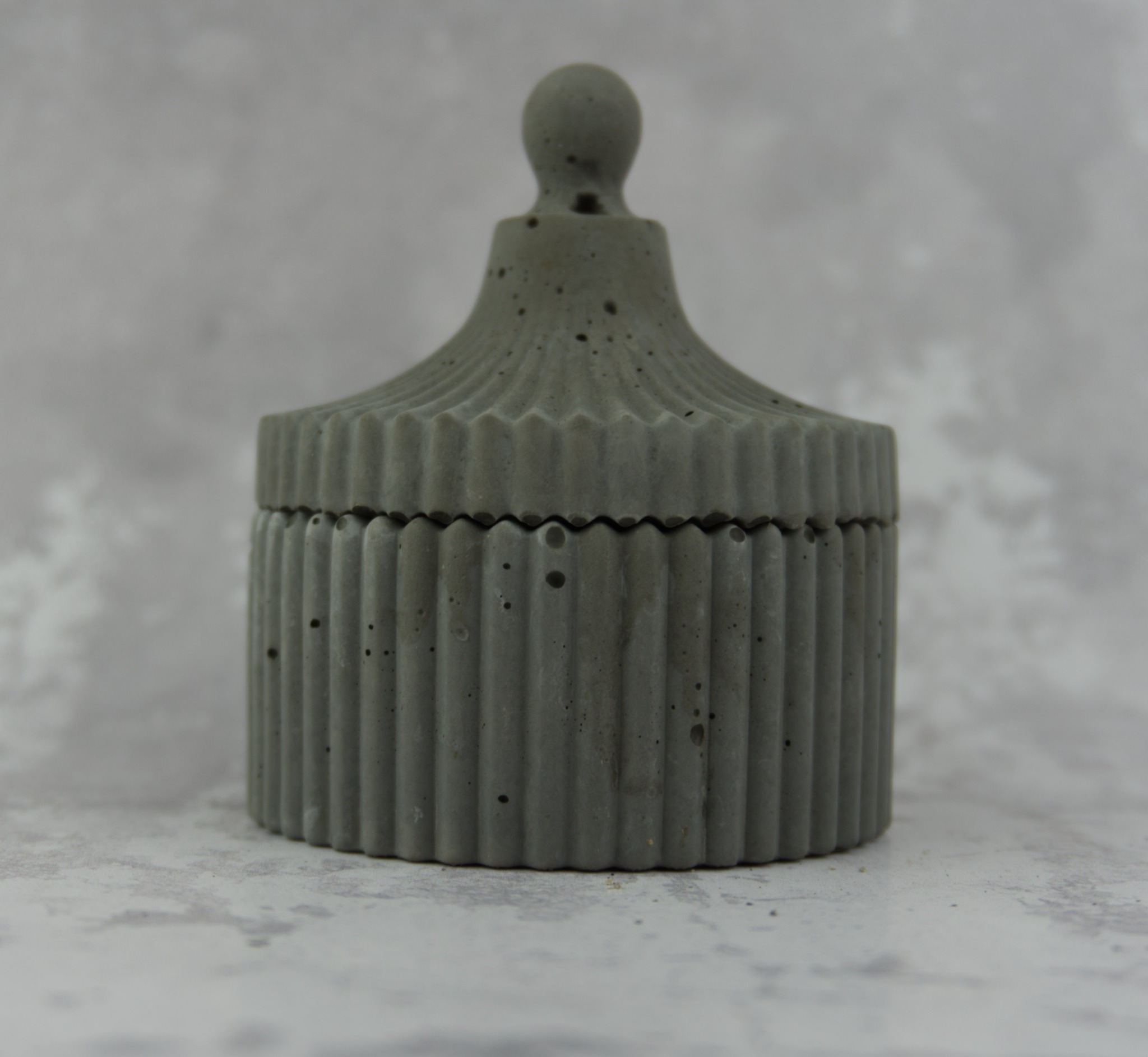 Szara, betonowa szkatułka z pokrywką i dekoracyjnymi pionowymi żłobieniami, na tle jasnej, marmurkowej powierzchni.