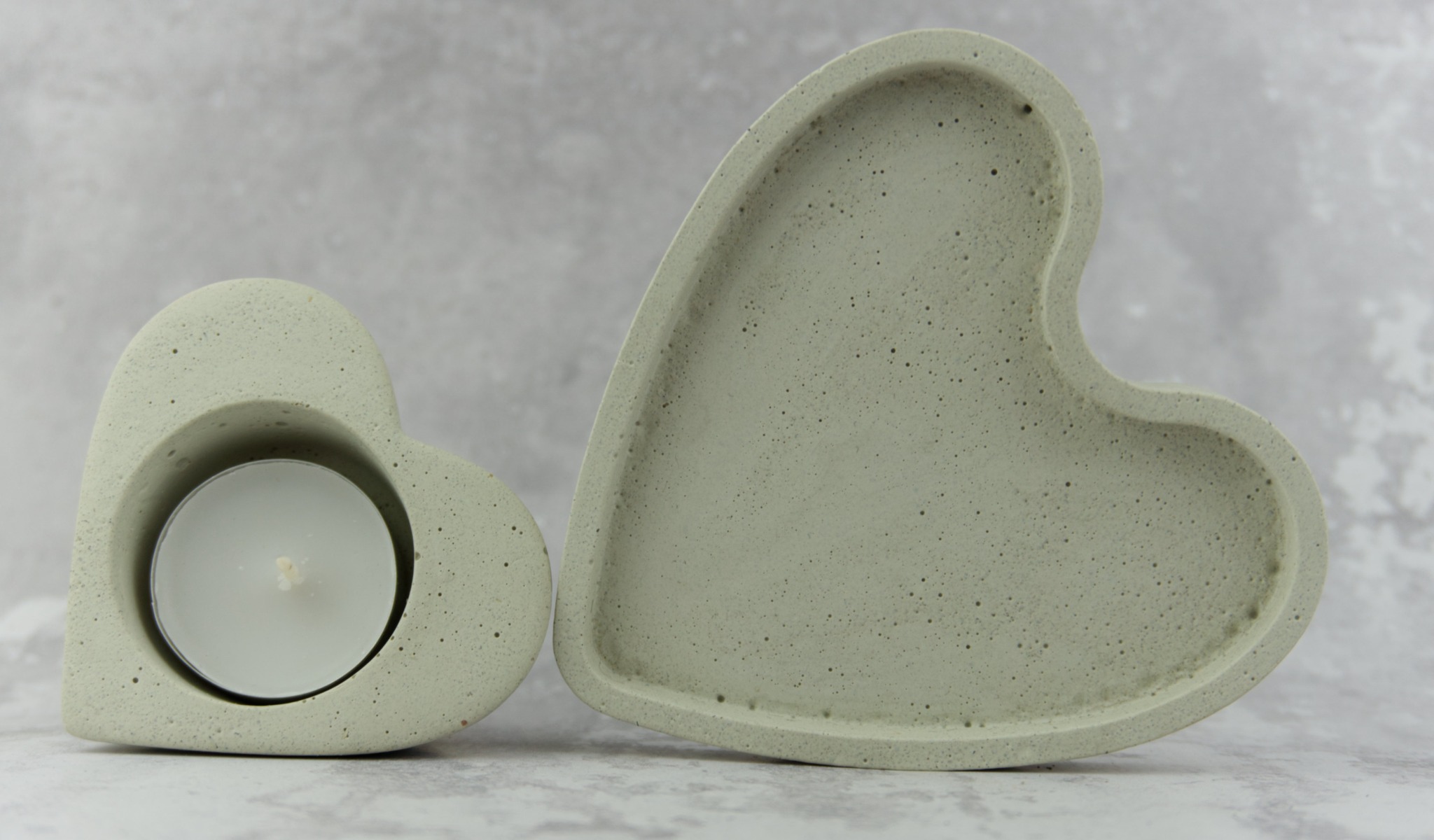 Dwa betonowe, szare serca: jedno puste, drugie z białą świeczką typu tealight w środku, na tle szarego marmuru.