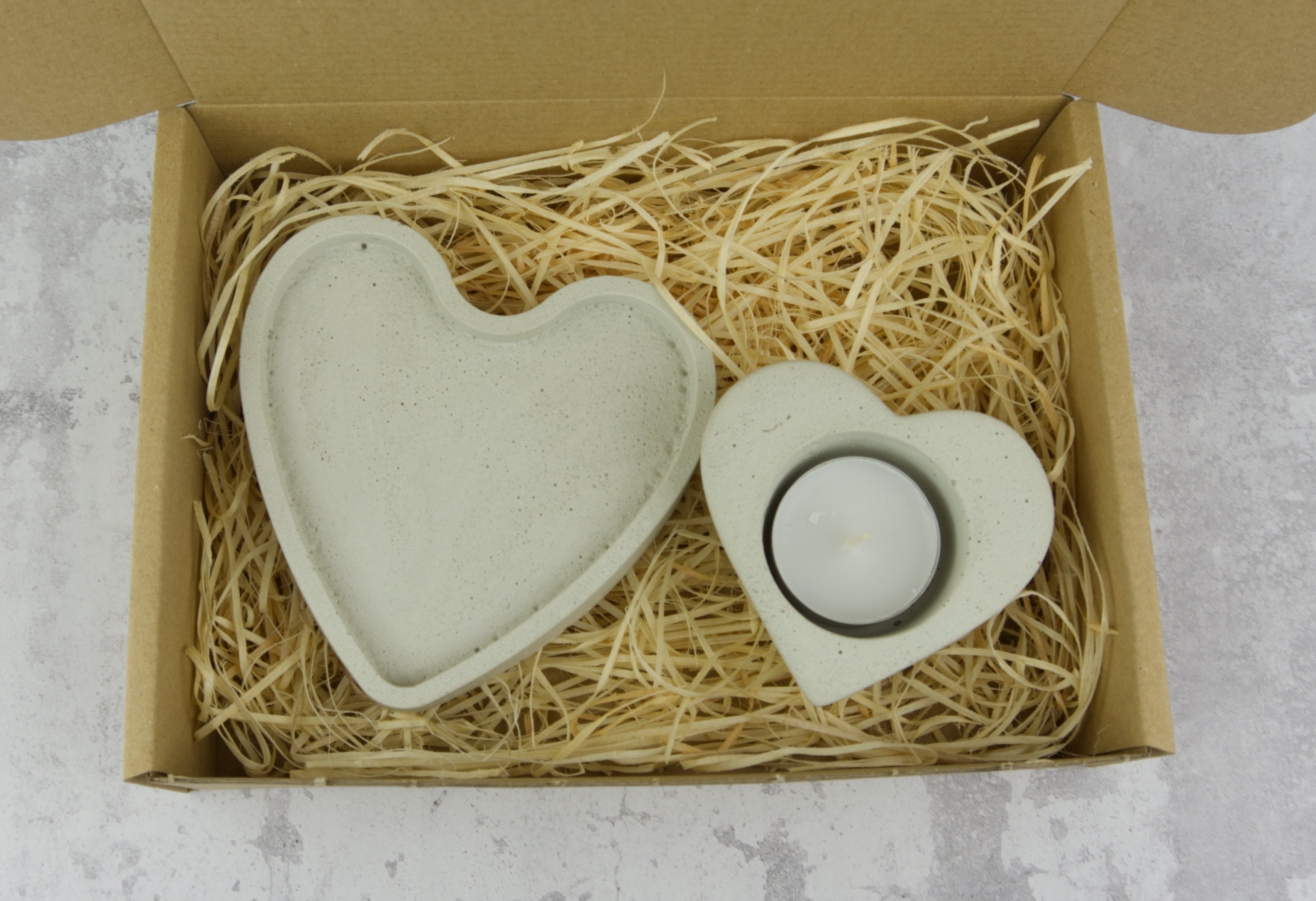 Dwa betonowe serca, jedno puste, drugie z białą świeczką typu tealight, ułożone na słomkowym wypełniaczu w kartonowym pudełku na tle szarej powierzchni.