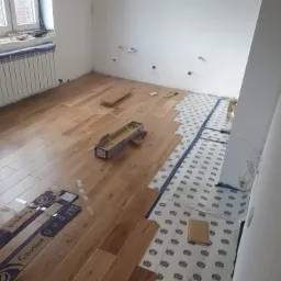 Proces układania jasnych paneli podłogowych w pokoju, widoczne paczki paneli Barlinek, podkład pod panele oraz fragmenty ułożonej podłogi.