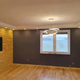 Jasne, przestronne wnętrze po metamorfozie: drewniana podłoga, ściana z dekoracyjnej cegły z telewizorem, szara ściana z oknem i nowoczesna lampa pierścieniowa.