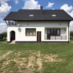 MICH-BUD - Dwukondygnacyjny dom murowany z ciemną dachówką, białymi ścianami i brązowymi ramami okiennymi, z balkonem i satelitą na bocznej ścianie, otoczony trawnikiem.