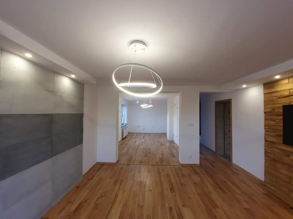 Nowoczesne wnętrze z drewnianą podłogą, ścianą z betonowych płyt i oświetleniem LED w kształcie pierścieni. Widoczne fragmenty ściany z drewnianą okładziną.