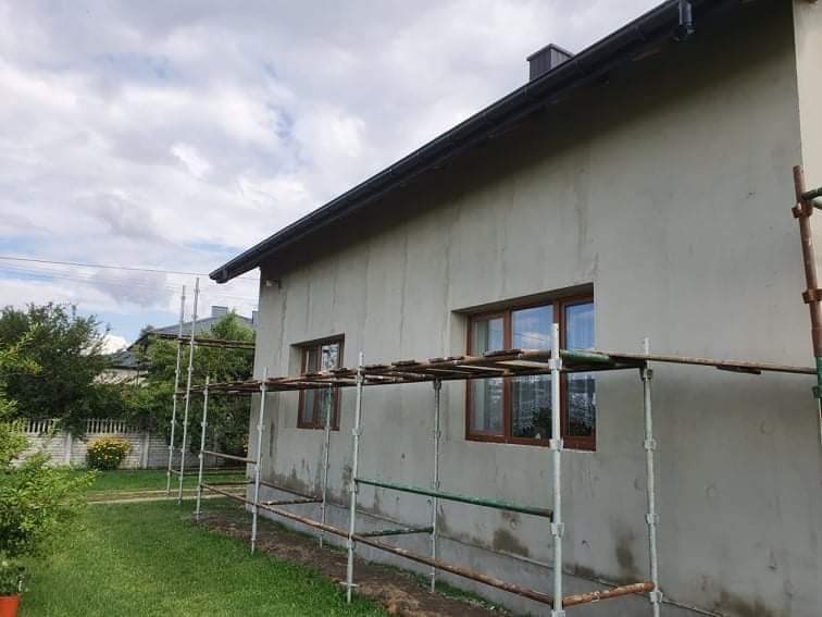 Ściana domu jednorodzinnego w trakcie tynkowania, widoczne rusztowanie, okna z brązowymi ramami i fragment ogrodu.