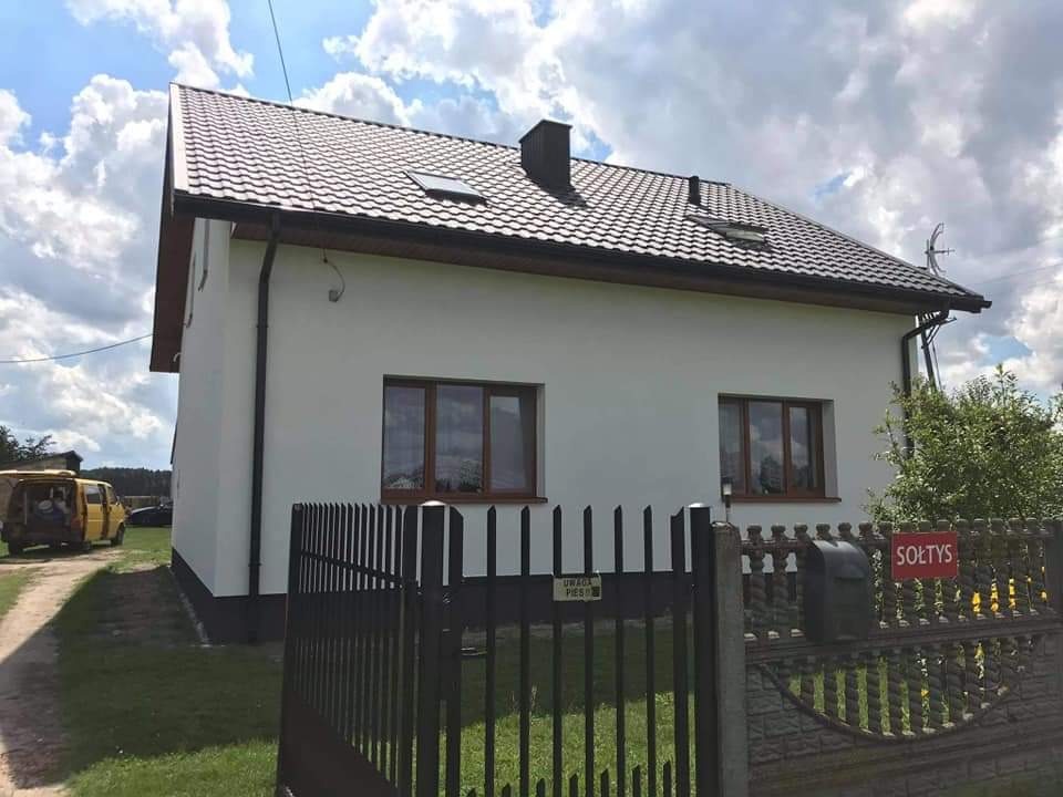 Nowo otynkowany, biały dom jednorodzinny z brązowym dachem i oknami, widoczny zza czarnego, metalowego ogrodzenia z tabliczką 'Sołtys'.
