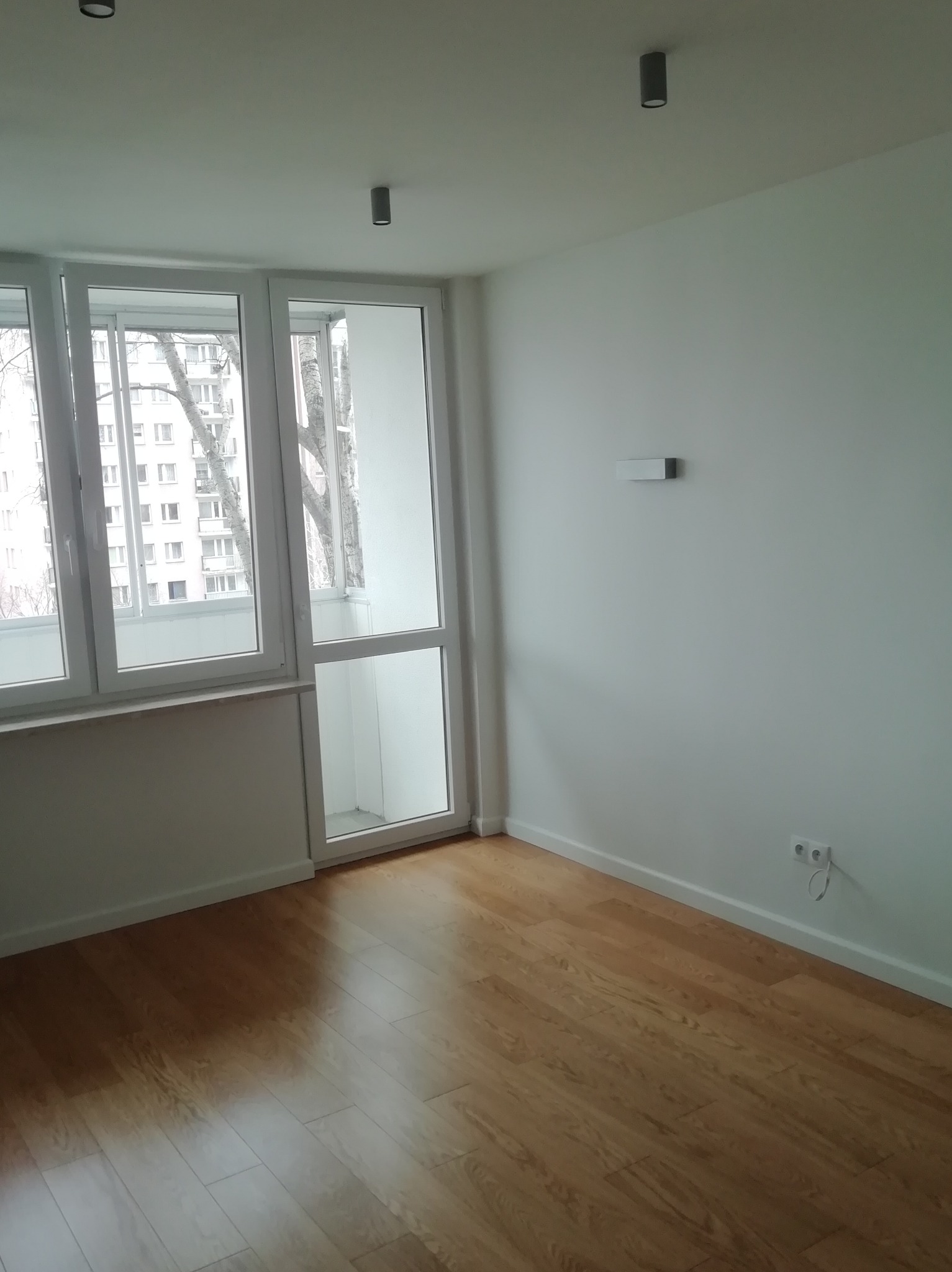 Pusty pokój po remoncie z jasnymi panelami podłogowymi, białymi ścianami i oknem balkonowym z widokiem na blok mieszkalny.