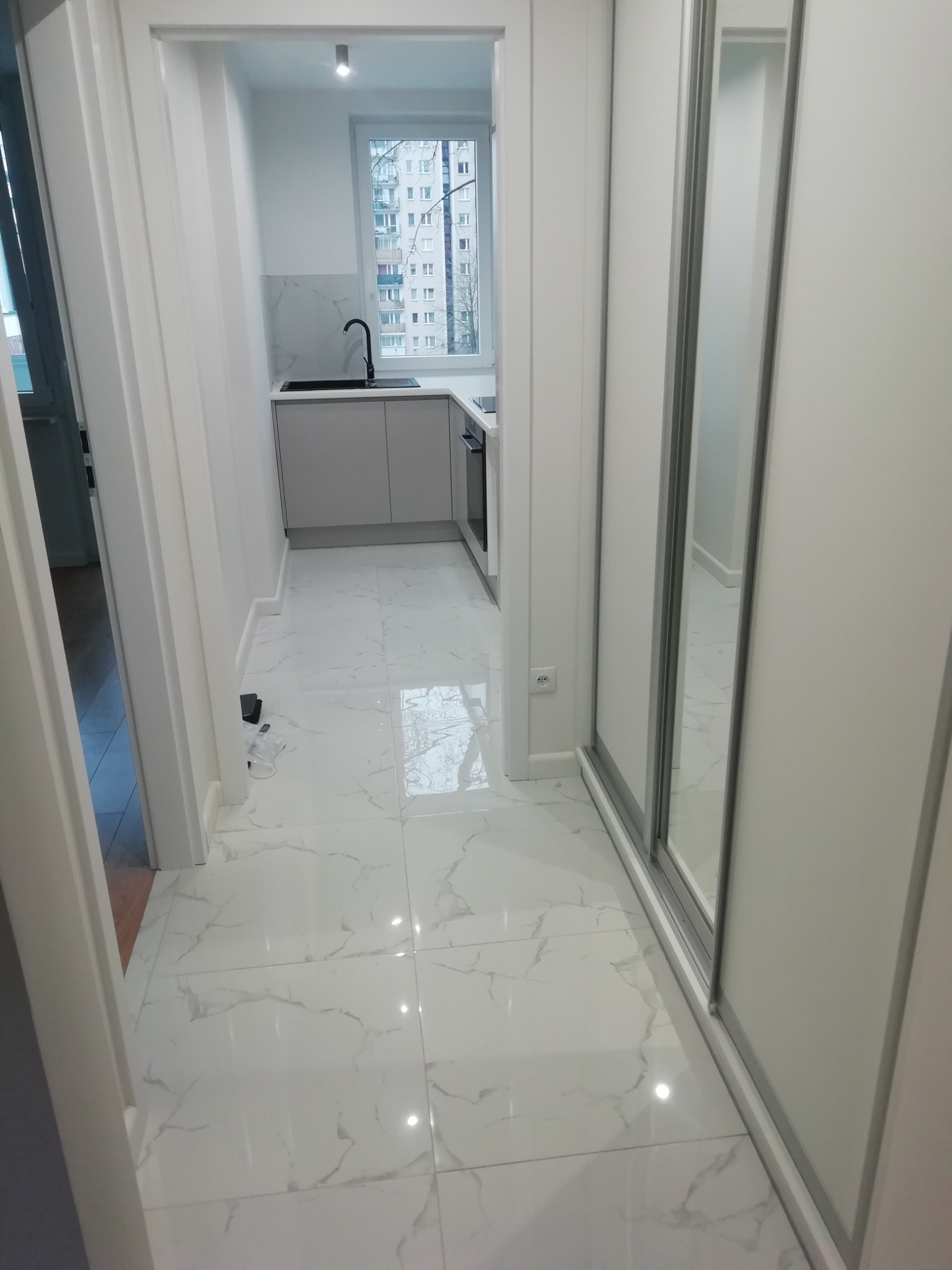 Jasne wnętrze mieszkania w Warszawie z białymi marmurowymi płytkami podłogowymi, widok na minimalistyczną kuchnię z czarną armaturą i szafą z przesuwnymi lustrzanymi drzwiami.