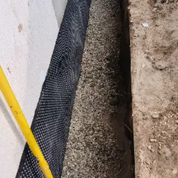 Izolacja fundament&oacute;w z folią kubełkową i drenażem żwirowym, widoczny wykop i ż&oacute;łty przew&oacute;d w pobliżu ściany fundamentowej.