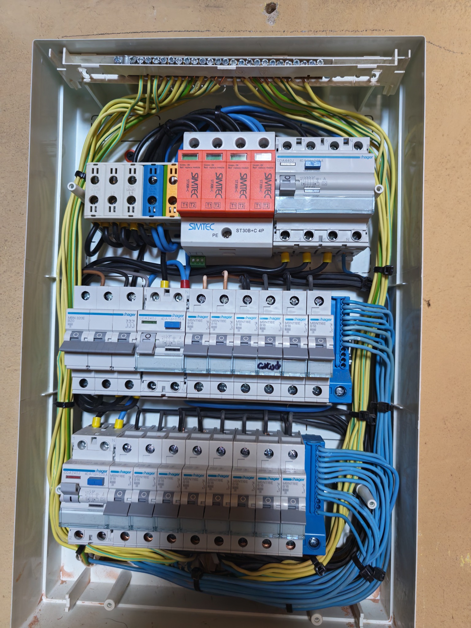Rozdzielnica elektryczna z zainstalowanymi bezpiecznikami, przewodami w kolorach żółtym, niebieskim i czarnym. Widoczne zabezpieczenia przeciwprzepięciowe.