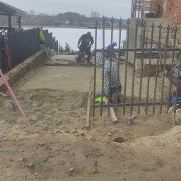 MASTER-BRUK HENRYK MATWIEJCZYK - Montaż metalowego ogrodzenia z ozdobnymi grotami na tle jeziora, widoczne narzędzia i prace ziemne w toku.