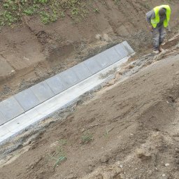 MASTER-BRUK HENRYK MATWIEJCZYK - Montaż krawężnika betonowego w rowie przydrożnym, pracownik w odblaskowej kamizelce zagęszcza podłoże ubijakiem, widoczne nierówności terenu i roślinność na skarpie.