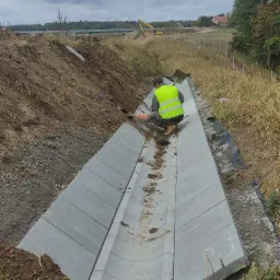 Montaż betonowego koryta odwadniającego na placu budowy, pracownik w żółtej kamizelce poziomuje elementy, w tle koparka i pochmurne niebo.