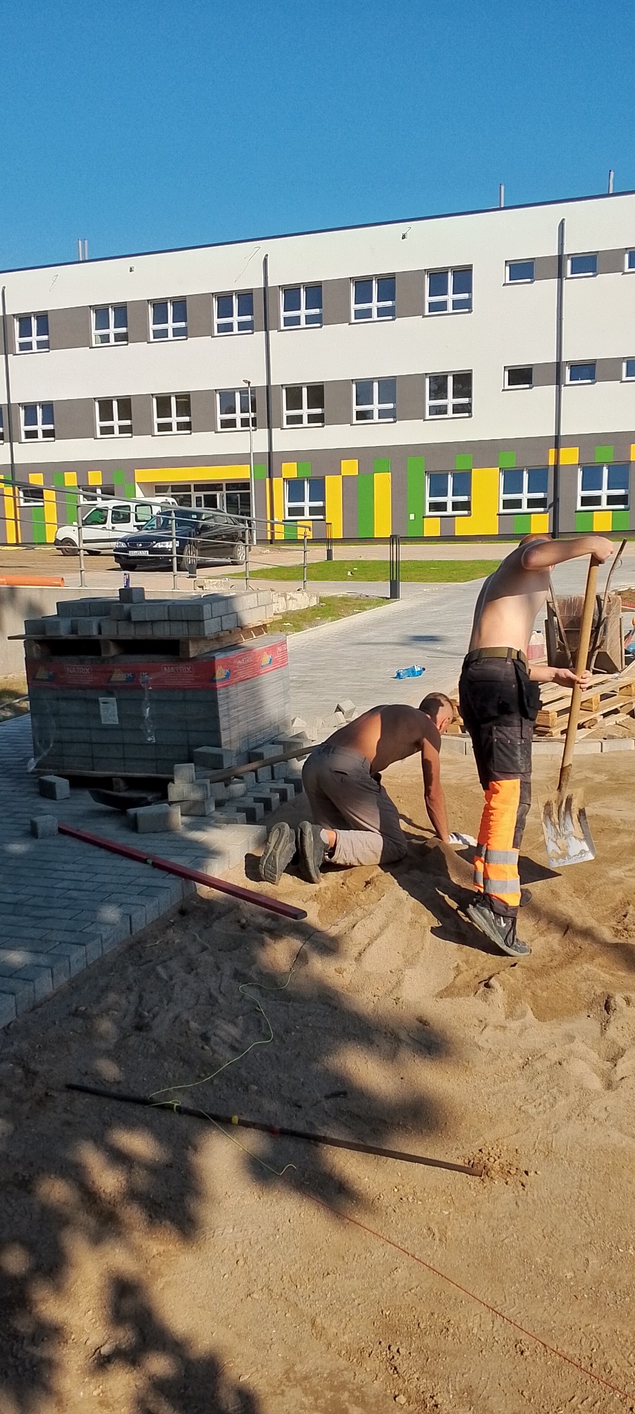 Dwóch robotników, jeden klęczy, drugi stoi z łopatą, układają kostkę brukową na piasku przed budynkiem z kolorowymi elementami, paleta kostki w tle.